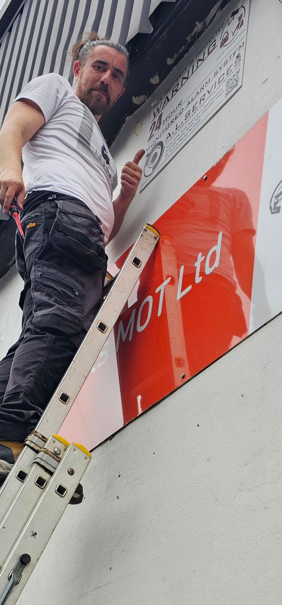 Andy Lord of ALServices up a ladder fitting a monitored alarm warning sign at a Lancashire MOT garage